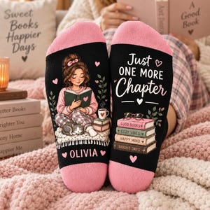 May include: A pair of black socks with pink cuffs featuring a graphic of a person reading a book with a cat, the name OLIVIA, and the text Just ONE MORE Chapter. The design includes a stack of books labelled Good Books, Cozy Vibes, Happy Mind, Bright Days.