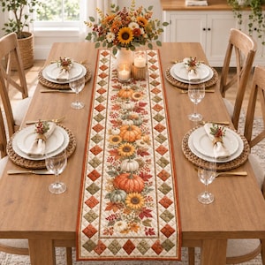 May include: A rectangular fabric table runner featuring a central print of orange pumpkins, sunflowers, and autumn leaves, bordered by a quilted diamond pattern in shades of orange, green, and cream, displayed on a wooden dining table with place settings.