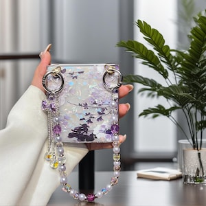 May include: A square compact mirror featuring an iridescent white surface with a purple floral pattern. The mirror is attached to a beaded strap made of light purple and clear beads, accented with a small purple bow charm and silver-toned hardware.