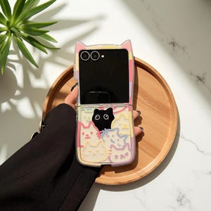 May include: A clear flip phone case featuring cat-shaped ears at the top and a colourful collage of various cat illustrations in pastel shades of pink, yellow, and blue, with one prominent black cat graphic centred on the lower half of the case.