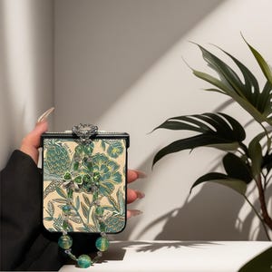 May include: A foldable smartphone case featuring a cream-colored background with a green and teal botanical floral pattern. The case includes a decorative beaded strap with green glass beads and a small green bow charm attached to the top.