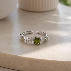 May include: A silver-toned open ring featuring a central square-cut olive green gemstone flanked by smaller clear round stones and additional tiny olive green accents along the band, creating a delicate and symmetrical arrangement of sparkling gems.