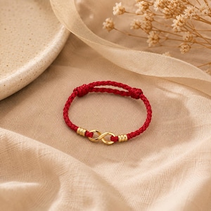 May include: A red braided cord bracelet featuring a central gold-toned infinity symbol charm. The infinity charm is flanked by two small gold-toned cylindrical beads on each side, securing the cord ends within the braided structure.