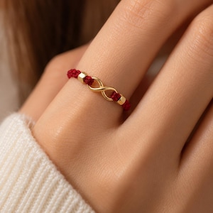 Puede incluir: Un anillo que presenta una banda de cordón rojo fino acentuada con un símbolo de infinito dorado central y dos pequeñas cuentas cilíndricas doradas colocadas a cada lado del dije, creando un diseño delicado y continuo.