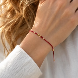 May include: A thin red cord bracelet featuring a small gold-toned infinity symbol charm at the center. The bracelet includes two tiny gold-toned beads flanking the infinity charm and two gold-toned beads at the adjustable cord ends.
