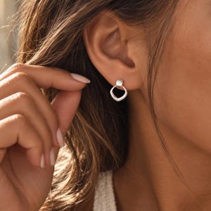 Pode incluir: Um pequeno brinco de tom prateado com um pendente liso em forma de quadrado arredondado, suspenso por um simples pino quadrado. O metal tem um acabamento polido que reflete a luz, criando uma aparência limpa e discreta no lóbulo da orelha.