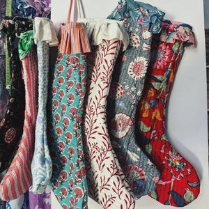 May include: A collection of six quilted Christmas stockings hanging in a row. Each stocking features a unique block-printed cotton fabric with floral or striped patterns, ruffled cuffs, and a hanging loop at the top in various vibrant colours.