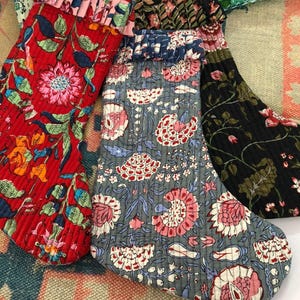 May include: Three quilted fabric stockings featuring floral patterns in red, grey, and black. Each stocking has a ruffled cuff at the top. The red stocking has pink and green flowers, the grey one has pink and white blooms, and the black one has floral vines.