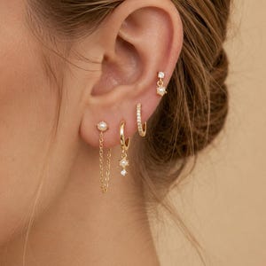 May include: A set of four gold earrings worn in the ear. One earring features a small pearl with a dangling chain. Another is a gold hoop with a pearl and crystal charm. A third is a thin gold hoop with crystals. The fourth is a small stud.