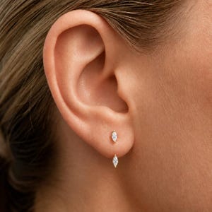 Puede incluir: Un pendiente de oro con dos piedras claras de talla marquesa dispuestas verticalmente. La piedra superior se asienta sobre el lóbulo, mientras que la inferior cuelga ligeramente por debajo, creando un aspecto delicado y alargado.
