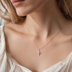 May include: A gold chain necklace featuring a cross pendant adorned with small white pearls and a single small pink gemstone. The delicate metal chain rests against the neck, showcasing the cross design with its pearl and gemstone accents.
