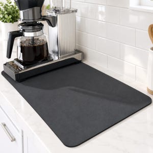May include: A rectangular, solid dark gray kitchen mat with rounded corners. The mat has a smooth, matte surface texture and is positioned flat on a countertop, partially underneath a black and clear glass coffee maker with a full carafe.
