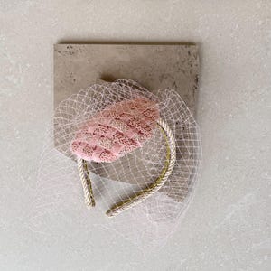 May include: A pink headband featuring a textured, woven fabric top piece adorned with a delicate, fine-mesh netting veil that drapes over the sides. The headband band is wrapped in a metallic gold-toned cord, providing a structured base.