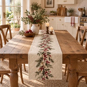 May include: A quilted beige table runner featuring a central botanical print of green pine branches, white berries, and red holly berries. The runner is centred on a rustic wooden dining table with two wooden chairs and a ceramic pitcher.