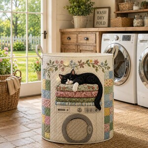 May include: A cylindrical laundry hamper featuring a quilted patchwork design in pastel colours. The front displays an embroidered graphic of a black and white cat sleeping on a stack of folded laundry above a depiction of a washing machine door.