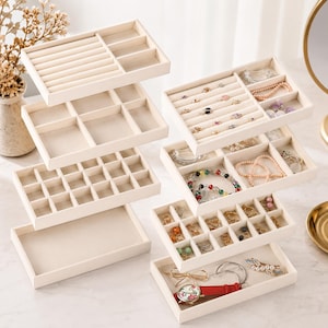 May include: A set of stackable, beige velvet-lined jewelry organizer trays. The trays feature various compartments, including ring rolls, small square grids, and open rectangular sections, some containing assorted necklaces, bracelets, and earrings.