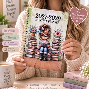 May include: A 2027-2029 monthly planner with a spiral binding. The cover features an illustration of a person with glasses and a blue hair bow sitting between two stacks of books, surrounded by flowers. The text reads 2027-2029 MONTHLY PLANNER.