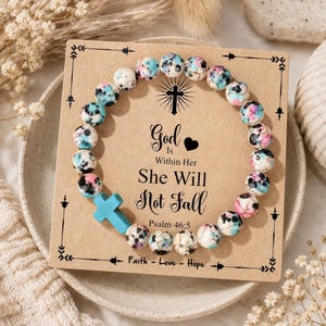 May include: A circular beaded bracelet featuring multi-coloured speckled beads in shades of blue, pink, and white, accented by a solid turquoise cross charm. The bracelet is displayed on a square tan card with the text God Is Within Her She Will Not Fall Psalm 46:5 and Faith - Love - Hope.