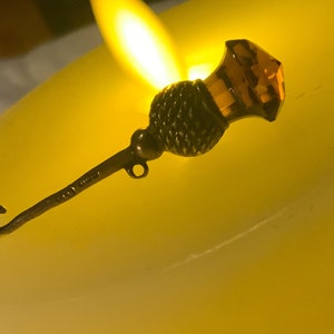 May include: A silver and amber colored thimble with a hook on the end. The thimble is sitting on a white surface.