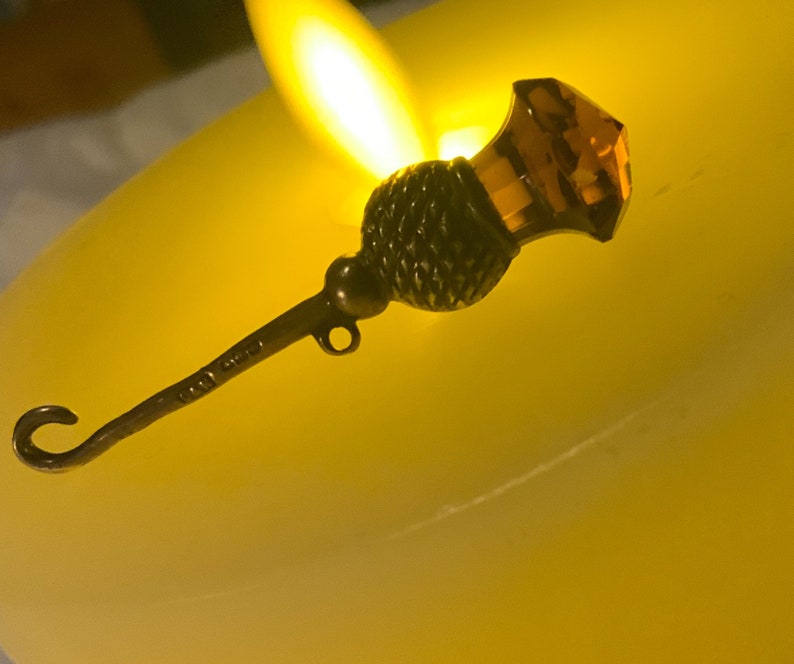 May include: A silver and amber colored thimble with a hook on the end. The thimble is sitting on a white surface.