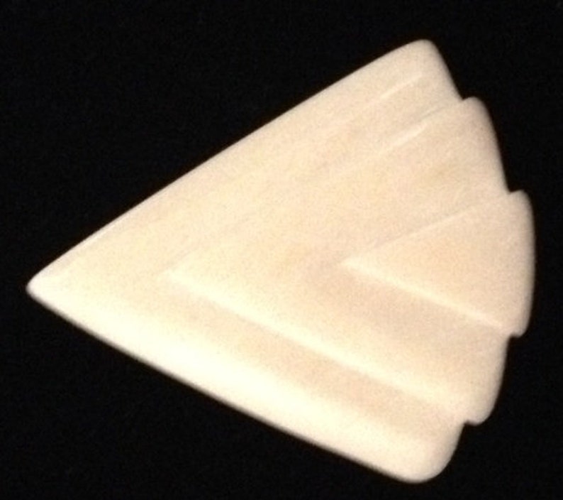 Op de afbeelding: Een witte, cr&egrave;mekleurige, keramische gitaarplectrum met een geometrisch ontwerp. De plectrum is driehoekig met afgeronde randen.