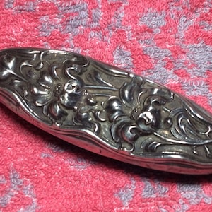 May include: A silver-toned oval-shaped trinket box with an ornate floral design. The box has a hinged lid and is decorated with a raised floral pattern.
