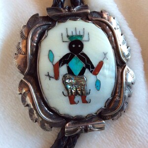 May include: A silver bolo tie with a round, white, and turquoise inlay featuring a stylized Native American figure. The figure is black with red and turquoise accents. The bolo tie is on a brown leather cord.
