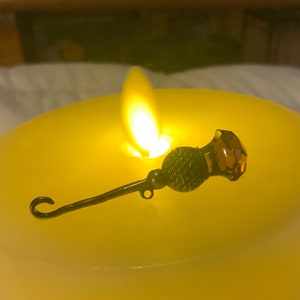 May include: A gold-toned metal pin with a dark brown thistle design and a clear amber glass top. The pin is sitting on a yellow candle.