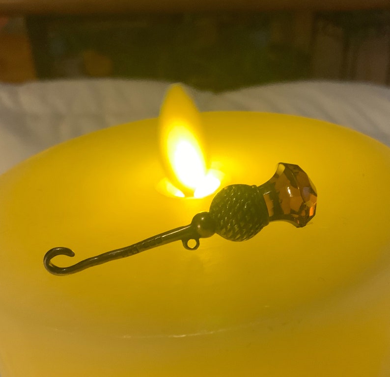 May include: A gold-toned metal pin with a dark brown thistle design and a clear amber glass top. The pin is sitting on a yellow candle.