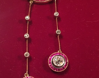 May include: A gold-toned necklace with a pendant featuring a diamond-studded oval and two dangling strands. Each strand has a series of diamonds and ends with a large, round pendant with a central diamond surrounded by red gemstones. The necklace is displayed on a red surface.
