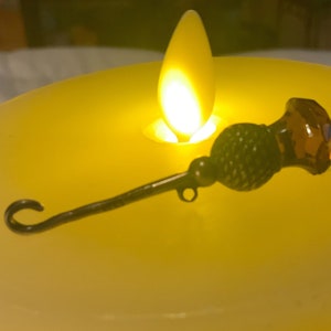 May include: A black metal and brown glass decorative pin with a looped end. The pin is sitting on a yellow candle.