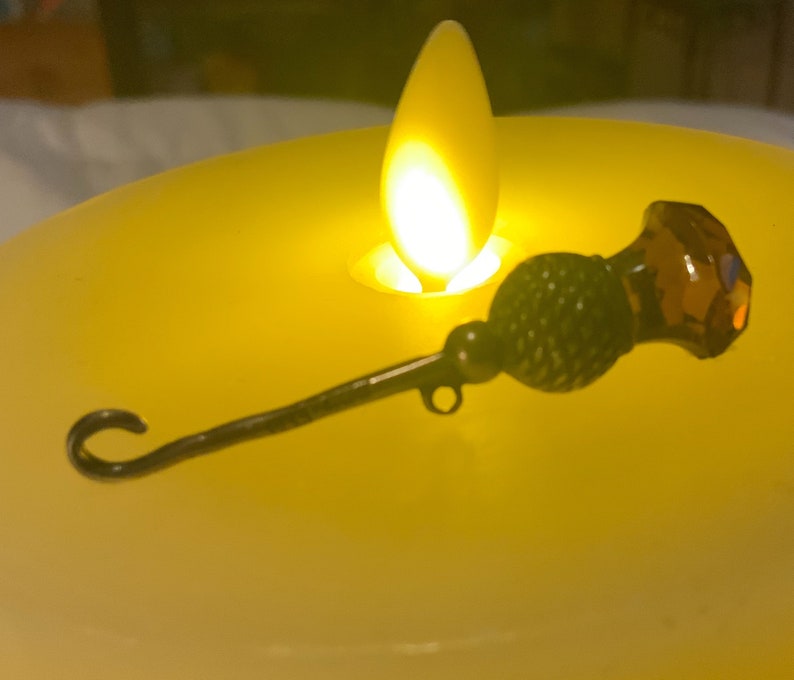 May include: A black metal and brown glass decorative pin with a looped end. The pin is sitting on a yellow candle.