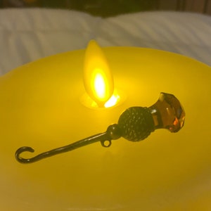 May include: A small, ornate, metal and glass, decorative object with a hook on one end and a faceted amber glass orb on the other. The object is sitting on a yellow candle.