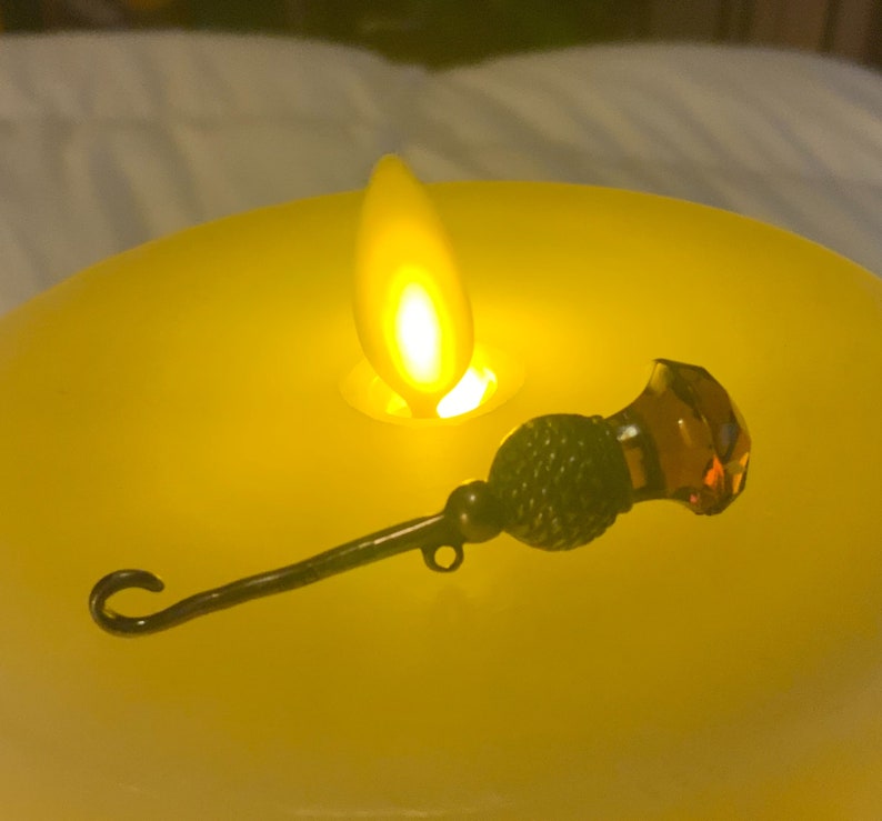 May include: A small, ornate, metal and glass, decorative object with a hook on one end and a faceted amber glass orb on the other. The object is sitting on a yellow candle.