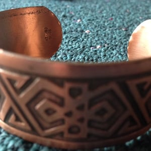 May include: A copper bracelet with a geometric pattern etched into the surface. The bracelet is stamped with the words "Solid Copper".