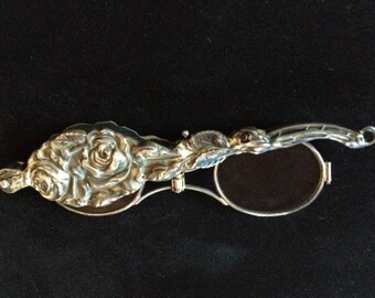 May include: A silver-toned magnifying glass with a rose design. The magnifying glass is closed and the rose design is visible on the front.