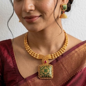 Puede incluir: Un conjunto de collar y pendientes jhumka en tono dorado. El collar presenta una cadena de tres hilos de cuentas con un gran colgante cuadrado que contiene esmalte floral verde y dorado y una pequeña piedra roja. Los pendientes son a juego.