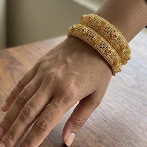 Puede incluir: Dos brazaletes dorados con un intrincado patrón de malla de filigrana, motivos circulares en relieve y pequeñas piedras rojas incrustadas. Las pulseras tienen un acabado mate texturizado y se usan juntas en la muñeca.
