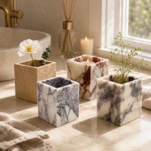 May include: Four square stone containers arranged on a surface. One is beige travertine, one is white with dark gray veining, one is white with red-brown veining containing a lit candle, and one is white with dark gray veining holding a green plant.