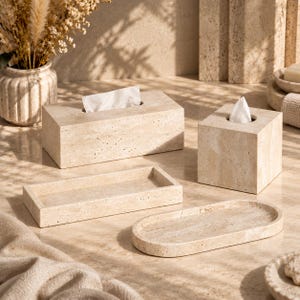 Puede incluir: Un conjunto de accesorios de baño de travertino beige que incluye una funda rectangular para caja de pañuelos, una cuadrada, una bandeja rectangular y una bandeja ovalada. Cada pieza presenta la textura porosa natural de la piedra.