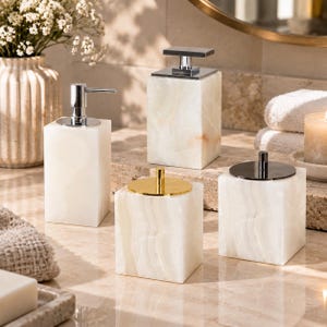 May include: A set of four white stone bathroom accessories featuring two rectangular soap dispensers with silver pumps and two square lidded canisters, one topped with a gold-toned circular lid and the other with a dark metallic circular lid.