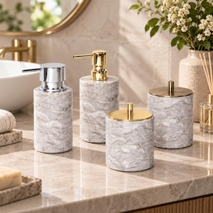 Puede incluir: Un juego de accesorios de baño compuesto por cuatro recipientes cilíndricos con un patrón de mármol gris y blanco. Dos recipientes tienen dispensadores de bomba, uno con acabado cromado y otro dorado. Dos recipientes tienen tapas con pomos dorados.