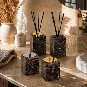 Puede incluir: Un juego de cuatro accesorios de baño cuadrados hechos de mármol marrón oscuro con vetas blancas. Dos artículos son difusores de varillas con palitos negros y bases doradas, y los otros dos son recipientes con tapas plateada y dorada.