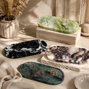May include: Four oval stone trays in various colours and patterns. One is black with white veining, one is green with dark streaks, one is white with deep purple marbling, and one is light green with subtle veining. Each tray has a smooth, polished finish.