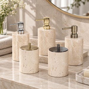 Puede incluir: Un conjunto de cinco accesorios de baño cilíndricos hechos de piedra travertino beige. Tres artículos son dispensadores de jabón con bombas en acabados de cromo pulido, oro y latón antiguo. Dos artículos son recipientes con tapas planas.