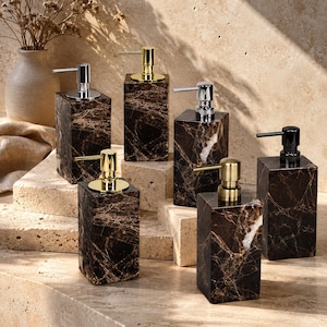 May include: Five rectangular soap dispensers made of dark brown marble with white veining. Each dispenser features a pump mechanism in varying metallic finishes, including polished chrome, brushed gold, and matte black, arranged on stone blocks.