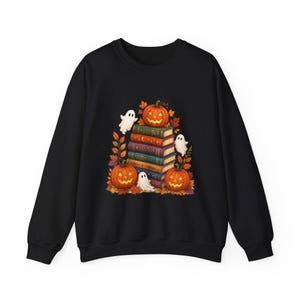 May include: A black long-sleeve sweatshirt featuring a central graphic of a stack of five colorful books surrounded by three carved orange jack-o'-lanterns, three small white ghosts, and various autumn-colored fall leaves in shades of red and gold.