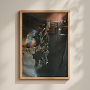 May include: A framed wall art print featuring a close-up of blue denim jeans with a brown leather belt. A silver metal carabiner is attached to a belt loop, holding a set of silver keys. A rainbow light refraction is visible on the denim fabric.
