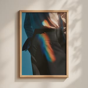 May include: A framed wall art print featuring a photograph of dark trousers and a light-coloured shirt. A vibrant, prismatic rainbow light flare stretches across the fabric, contrasting with the deep blue background and the wooden frame.