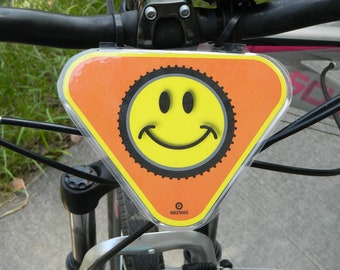 Może przedstawiać: Czarny rower z żółtą naklejką ze śmieszkiem na trójkątnym pomarańczowym znaku. Znak ma napis "Bike Lane" na dole.